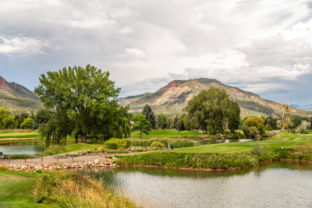 A scenic landscape photograph featuring a lush green dalton ranch golf course in durango colorado with a calm lake in the foreground, surrounded by trees and rugged mountains under a cloudy sky.